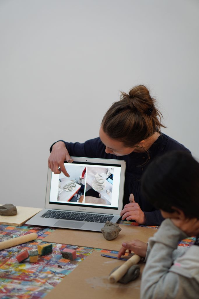 An artist pointing to a laptop screen which is showing a clay model to a group of children with modelling clay at a workshop inspired by Jagjit Kaur's work.